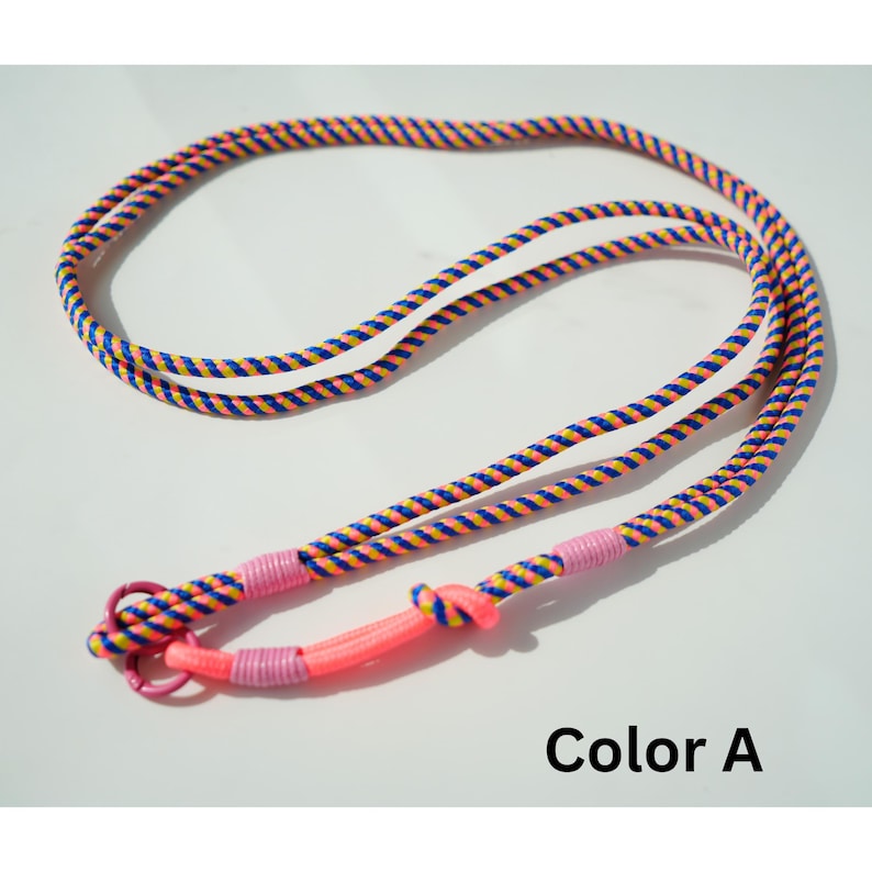 Pode incluir: Uma trela de corda comprida com um padr&atilde;o torcido de fios azuis, amarelos e cor-de-rosa. A trela inclui um anel met&aacute;lico cor-de-rosa numa extremidade, fixado com enrolamentos de cord&atilde;o rosa claro e um n&oacute; rosa n&eacute;on. Texto Color A vis&iacute;vel.
