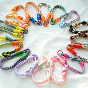 Puede incluir: Una colección de pulseras de cuerda coloridas en varios tonos, incluyendo naranja, amarillo, verde, morado y rosa. Cada pulsera presenta un diseño de nudo único y está acentuada con herrajes de plástico o metal a juego. Las pulseras están dispuestas en un patrón circular.