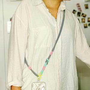 Pode incluir: Um cord&atilde;o de corda com padr&atilde;o cinza e branco com detalhes em rosa e verde menta, um anel conector amarelo brilhante e um acess&oacute;rio de capa transparente para celular, usado diagonalmente sobre uma camisa branca.