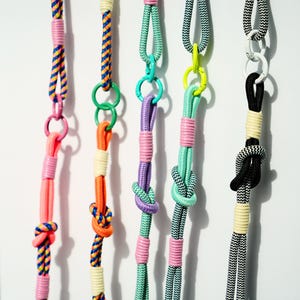 Pode incluir: Cinco cord&otilde;es verticais feitos de corda de nylon tran&ccedil;ada em v&aacute;rias cores, incluindo rosa neon, laranja, roxo, verde menta e padr&otilde;es em preto e branco. Cada cord&atilde;o apresenta an&eacute;is de metal, n&oacute;s decorativos e detalhes enrolados.