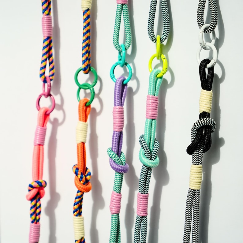 Pode incluir: Cinco cord&otilde;es verticais feitos de corda de nylon tran&ccedil;ada em v&aacute;rias cores, incluindo rosa neon, laranja, roxo, verde menta e padr&otilde;es em preto e branco. Cada cord&atilde;o apresenta an&eacute;is de metal, n&oacute;s decorativos e detalhes enrolados.