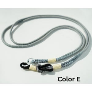 Pode incluir: Um cord&atilde;o longo com padr&atilde;o de chevron cinza e branco, apresentando uma se&ccedil;&atilde;o de corda com padr&atilde;o de chevron preto e branco amarrada em um n&oacute;. A corda &eacute; fixada com fios de cor creme e inclui um anel de metal branco. O texto diz Color E.