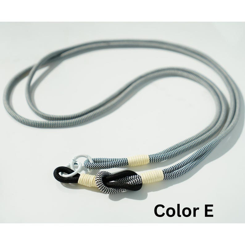 Pode incluir: Um cord&atilde;o longo com padr&atilde;o de chevron cinza e branco, apresentando uma se&ccedil;&atilde;o de corda com padr&atilde;o de chevron preto e branco amarrada em um n&oacute;. A corda &eacute; fixada com fios de cor creme e inclui um anel de metal branco. O texto diz Color E.