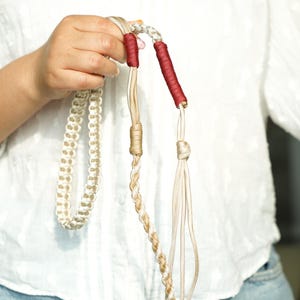 Op de afbeelding: Een handgemaakt macramé-accessoire met een reeks crèmekleurige geknoopte koorden, een gevlochten gedeelte en twee afzonderlijke segmenten omwikkeld met donkerrood garen. Het ontwerp bevat meerdere texturen en lussen, wat complexe knooptechnieken laat zien.