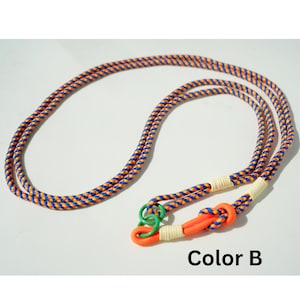 Pode incluir: Um cord&atilde;o longo de corda tran&ccedil;ada com um padr&atilde;o repetitivo de fios azuis, laranja e amarelos. A extremidade inclui um la&ccedil;o laranja brilhante, um pequeno anel verde e dois enrolamentos de corda cor de creme. O texto Color B est&aacute; impresso abaixo.