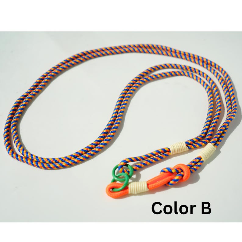 Pode incluir: Um cord&atilde;o longo de corda tran&ccedil;ada com um padr&atilde;o repetitivo de fios azuis, laranja e amarelos. A extremidade inclui um la&ccedil;o laranja brilhante, um pequeno anel verde e dois enrolamentos de corda cor de creme. O texto Color B est&aacute; impresso abaixo.