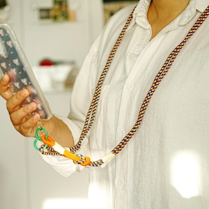 Pode incluir: Um cord&atilde;o longo para telem&oacute;vel com uma corda entran&ccedil;ada em azul-marinho, amarelo e branco. A al&ccedil;a inclui um cursor ajust&aacute;vel laranja brilhante, um anel de pl&aacute;stico verde e uma capa transparente com um padr&atilde;o floral branco discreto.