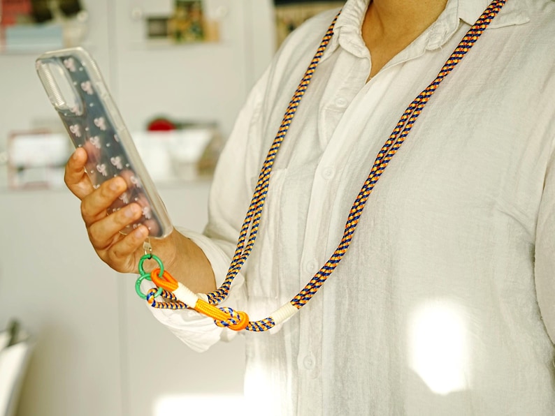 Pode incluir: Um cord&atilde;o longo para telem&oacute;vel com uma corda entran&ccedil;ada em azul-marinho, amarelo e branco. A al&ccedil;a inclui um cursor ajust&aacute;vel laranja brilhante, um anel de pl&aacute;stico verde e uma capa transparente com um padr&atilde;o floral branco discreto.