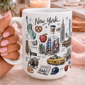 May include: A white ceramic mug featuring a colorful watercolor-style illustration of New York City landmarks and icons, including the Statue of Liberty, a yellow taxi, the Empire State Building, a pretzel, an apple, and the text New York and NYC.