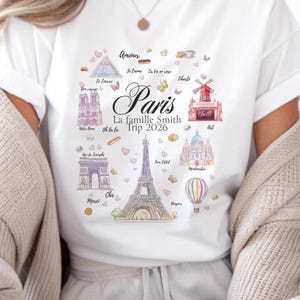 May include: White t-shirt featuring a colorful watercolor design of Paris landmarks, including the Eiffel Tower, Arc de Triomphe, and Moulin Rouge. The shirt includes the text "Paris La famille Smith Trip 2026" and other French phrases.