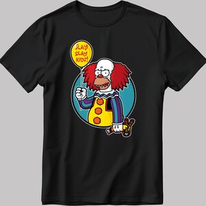May include: Black t-shirt featuring a cartoon illustration of a clown holding a red balloon that reads "SLAY SLAY KIDS!" The clown is wearing a yellow, purple and blue striped shirt with a red bow tie and red hair. The clown is holding a slingshot in its other hand.