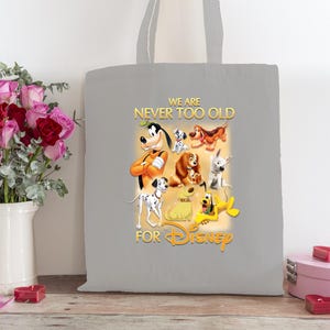 Peut inclure: Un sac fourre-tout gris avec un graphisme coloré de chiens de dessins animés Disney, dont Pluto, Dingo et Lady. Le texte "WE ARE NEVER TOO OLD FOR Disney" est imprimé en or. Un bouquet de roses est dans un vase blanc.