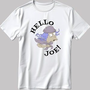 Op de afbeelding: Wit T-shirt met een cartoon afbeelding van een vrouw met blauw haar en de tekst "HELLO JOE!" gedrukt op de voorkant.