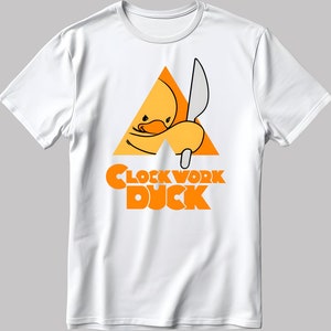 May include: White t-shirt featuring an orange graphic of a duck holding a knife within a triangle. The text "Clockwork Duck" is positioned beneath the graphic.