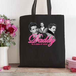 Può includere: Borsa tote nera con un'immagine in bianco e nero di un uomo e il testo rosa "Daddy is a state of mind". Un mazzo di rose rosse e rosa è accanto alla borsa. La borsa ha un manico.