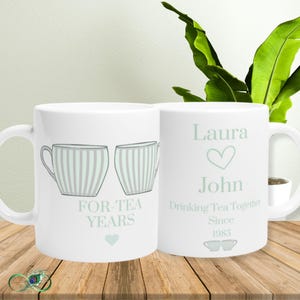 Peut inclure: Deux mugs en céramique blancs. L'un présente deux tasses à thé à rayures verticales et le texte "FOR-TEA YEARS". L'autre mug porte les noms "Laura" et "John", un cœur et le texte "Drinking Tea Together Since 1985".
