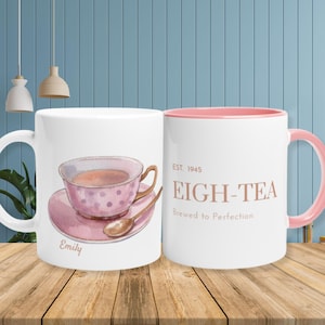 Könnte beinhalten: Weißer Keramikbecher mit rosa Innenraum und Henkel. Eine Seite zeigt eine Aquarellillustration einer Teetasse und Untertasse mit dem Namen "Emily". Die andere Seite hat den Text "EIGH-TEA" und "Brewed to Perfection".