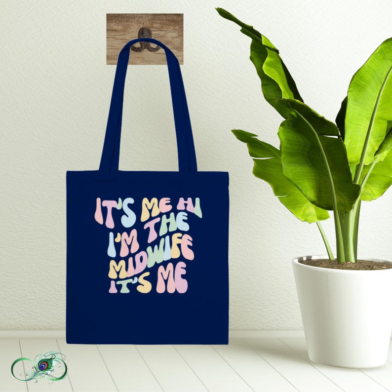 Op de afbeelding: Een donkerblauwe tote bag met de tekst "It's me, I'm the midwife, it's me" in een regenboogkleurig retro-lettertype.