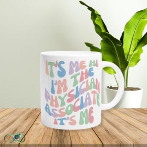 Könnte beinhalten: Weiße Keramiktasse mit einem bunten Design, auf dem "It's me hi I'm the Physician Associate It's me" steht.