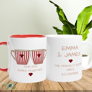 May include: Two white ceramic mugs with red interiors and handles. One mug features a graphic of two striped teacups with red gems and the text FOR-TEA YEARS TOGETHER. The other mug displays the text EMMA & JAMES THE PERFECT BLEND SINCE 20.08.1986.