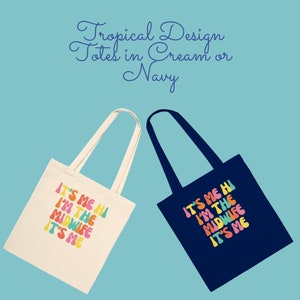 Op de afbeelding: Twee tote bags met de tekst "IT'S ME HI I'M THE MIDWIFE IT'S ME" in kleurrijke, retro-stijl letters. E&eacute;n tas is cr&egrave;mekleurig en de andere is marineblauw.