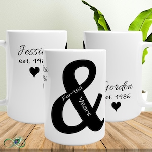 Puede incluir: Tres tazas de cerámica blanca con texto negro y detalles de corazón. Una taza presenta el nombre "Jessica" y el año "1986". Otra taza tiene el nombre "Gordon" y el año "1986". La tercera taza tiene una gran & con las palabras "For-tea Years".