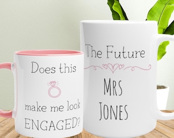 Op de afbeelding: Twee witte keramische mokken met zwarte tekst en roze accenten. Eén mok leest "Does this make me look ENGAGED?" met een ringafbeelding. De andere mok zegt "The Future MRS JONES".