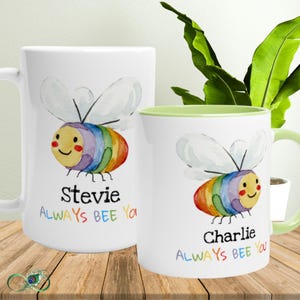 Può includere: Due tazze in ceramica bianca con un disegno ad acquerello di un'ape a strisce arcobaleno. Le tazze riportano la scritta "Stevie Always BEE You" e "Charlie Always BEE You" in un font colorato. L'interno di una tazza è verde.