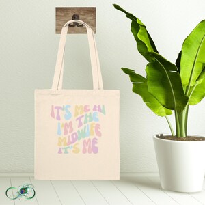 Op de afbeelding: Een beige tote bag met een kleurrijke afbeelding met de tekst "It's me, I'm the midwife, it's me".