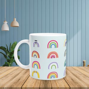 Op de afbeelding: Witte keramische mok met een lichtblauwe achtergrond versierd met kleurrijke regenboog illustraties. De regenboogjes variëren in grootte en kleur, wat zorgt voor een vrolijk ontwerp. De mok heeft een grote, gebogen handgreep.
