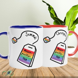 Op de afbeelding: Twee witte keramische mokken met blauwe en rode binnenkant en handvatten. Elke mok heeft een theezakje-ontwerp met de woorden "Neuro-positivi-TEA" in regenboogkleuren en een naam. Er is ook een regenbooghart aanwezig.