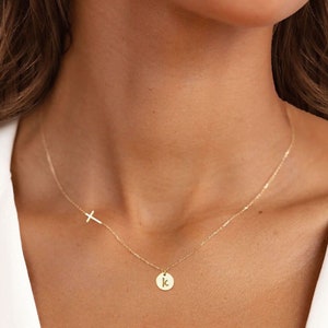 May include: A gold chain necklace with a small, round pendant featuring the letter "K" and a delicate cross charm.
