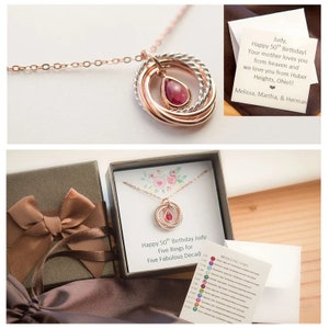 Op de afbeelding: Een gouden ketting met een hanger van vijf in elkaar grijpende ringen en een rode edelsteen. De ketting zit in een cadeaudoos met een kaart waarop staat "Happy 50th Birthday Judy Five Rings for Five Fabulous Decades".