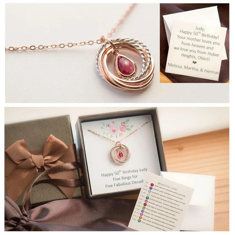 Op de afbeelding: Een gouden ketting met een hanger van vijf in elkaar grijpende ringen en een rode edelsteen. De ketting zit in een cadeaudoos met een kaart waarop staat "Happy 50th Birthday Judy Five Rings for Five Fabulous Decades".