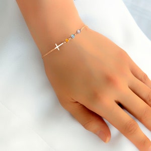 May include: A delicate gold bracelet featuring a cross charm and three colorful gemstone accents. The bracelet is shown on a wrist, showcasing its elegant design and subtle sparkle. The gemstones are yellow, blue, and pink.