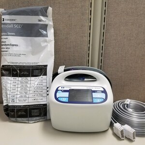 May include: A white medical device with a blue display and a handle. The device is labelled "Kendall SCD" and has a coiled hose attached. A white bag with a label that reads "Kendall SCD" and "Compression Sleeves" is in the background.