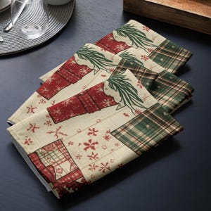 Könnte beinhalten: Ein Stapel von vier weihnachtlichen Stoffservietten. Jede Serviette hat einen cremefarbenen Hintergrund mit roten Geschenkboxen, Schneeflocken und grünen Karomustern. Die Servietten sind gefaltet und auf einer dunkelblauen Oberfläche angeordnet.