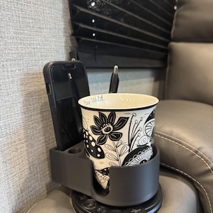 May include: A black and white floral-patterned coffee cup with the text "Stay Wild" sits in a gray cup holder with a phone slot. The cup holder is mounted on a gray leather couch, near a window with black blinds.