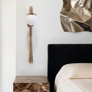 May include: A wall-mounted light fixture with a spherical white glass shade and a woven macrame design. The fixture has a brown tassel and is mounted on a white wall. A marble-patterned nightstand is below the light.