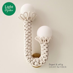 May include: A wall sconce featuring two white spherical glass globes held by a U-shaped metal frame wrapped in thick, cream-colored macrame cord. The cord is knotted in a repeating pattern, and the fixture is mounted on a circular brass base.