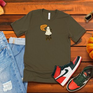 Pu&ograve; includere: Una maglietta verde scuro con una grafica di un fantasma bianco e il testo "Boo-dies" in arancione. Il fantasma ha quattro gambe e tiene una piccola zucca. La maglietta &egrave; su una superficie di legno con un paio di jeans, una zucca, occhiali da sole e scarpe da ginnastica.