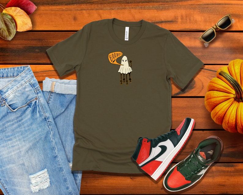 Pu&ograve; includere: Una maglietta verde scuro con una grafica di un fantasma bianco e il testo "Boo-dies" in arancione. Il fantasma ha quattro gambe e tiene una piccola zucca. La maglietta &egrave; su una superficie di legno con un paio di jeans, una zucca, occhiali da sole e scarpe da ginnastica.