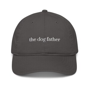 May include: A charcoal gray baseball cap with a curved brim. The text "the dog father" is embroidered in white across the front of the cap. The cap is made of a soft, durable material.