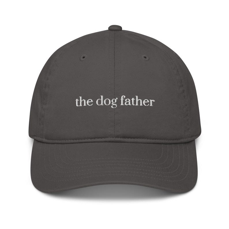 May include: A charcoal gray baseball cap with a curved brim. The text "the dog father" is embroidered in white across the front of the cap. The cap is made of a soft, durable material.