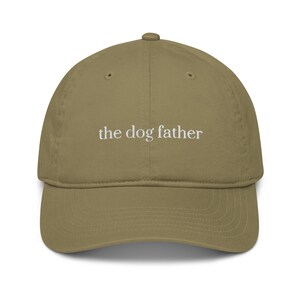 May include: A khaki-colored baseball cap with a curved brim. The text "the dog father" is embroidered in white across the front of the cap. The cap has a classic six-panel design and an adjustable strap.