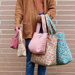 A person holding a collection of fabric tote bags in various sizes and floral patterns. The bags are in shades of pink, beige, and teal. The background is a brick wall.