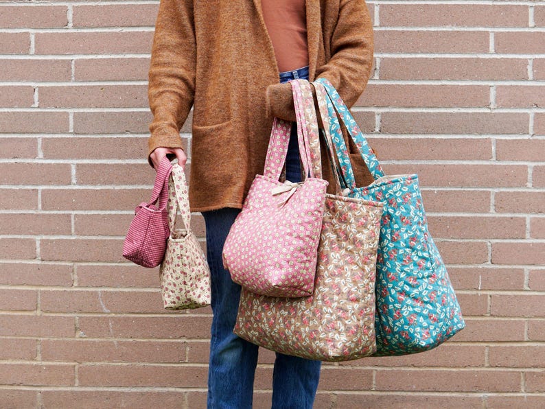 A person holding a collection of fabric tote bags in various sizes and floral patterns. The bags are in shades of pink, beige, and teal. The background is a brick wall.