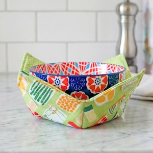 May include: A blue and white patterned ceramic bowl nested inside a green and patterned fabric bowl cozy. The cozy has a light green exterior with colorful food-related graphics. The bowl is on a white countertop.