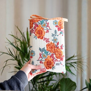 Puede incluir: Una funda rectangular con estampado floral, con fondo crema, con flores naranjas y azules y hojas verdes. El borde superior es naranja y una cinta a juego está atada en la parte superior. Las palabras "Milk & Honey" están impresas.