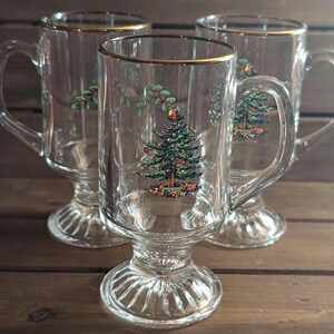 Op de afbeelding: Drie heldere glazen mokken met gouden randen en handvatten. Elke mok heeft een kerstboomontwerp met kleurrijke ornamenten. De mokken staan op een houten oppervlak.